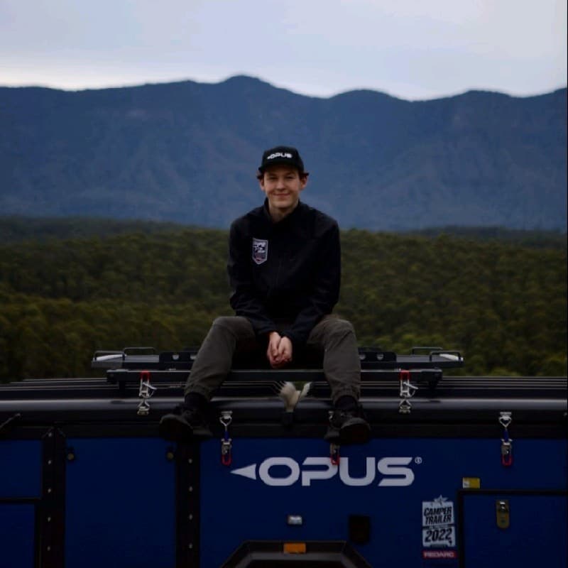 Joel Stocklinger sitting on an OPUS camper trailer, with forested ranges behind